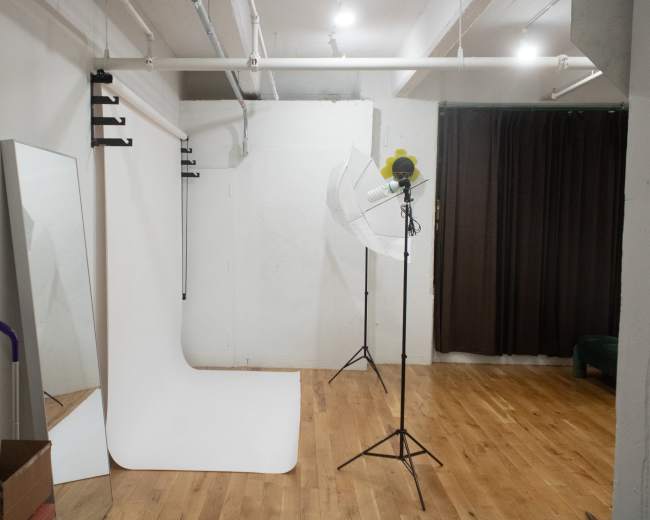 The image shows a photography studio with a white backdrop, a large light umbrella, and a mirror against a wall.