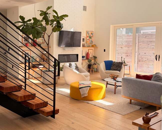 The image depicts a bright and spacious modern living room with a staircase, a sectional sofa, a round yellow ottoman, and large windows overlooking a patio area.