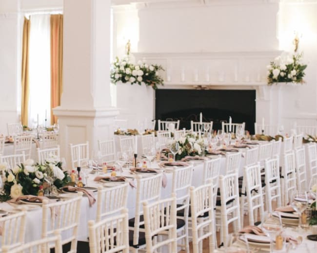 The image shows a banquet hall set for a formal dinner with white tables, chairs, and floral arrangements.