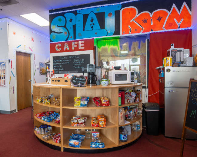 A cafe area with a round wooden shelf displaying snacks and beverages, surrounded by colorful wall decor and a service counter.