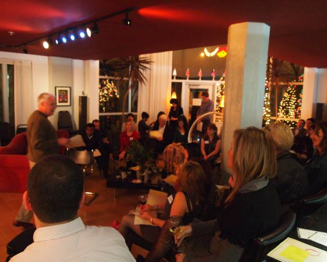 A group of people is seated in a well-lit indoor space, with a speaker presenting at the front and decorations including plants and lights in the background.