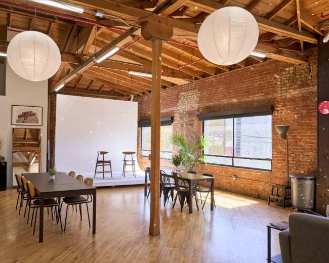 The image shows a spacious and well-lit interior with exposed brick walls, wooden beams, a seating area with tables and chairs, and a small stage at the back featuring two stools.