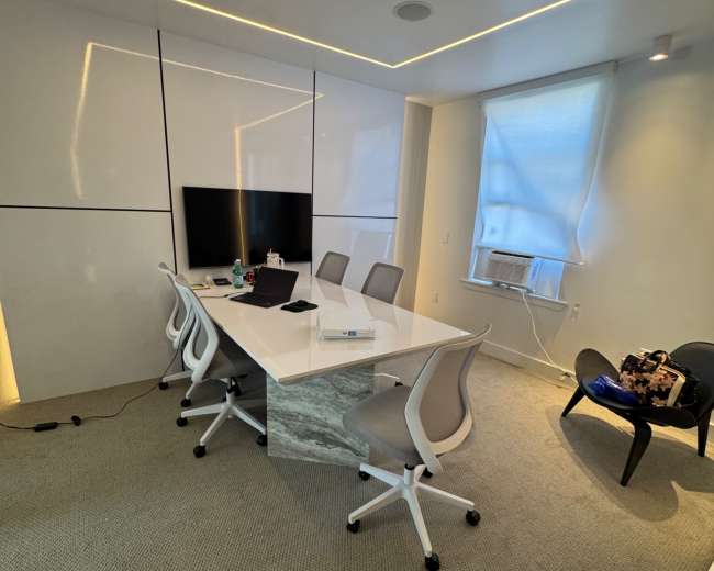 A modern conference room features a white table and chairs, a wall-mounted television, and a window with an air conditioning unit.