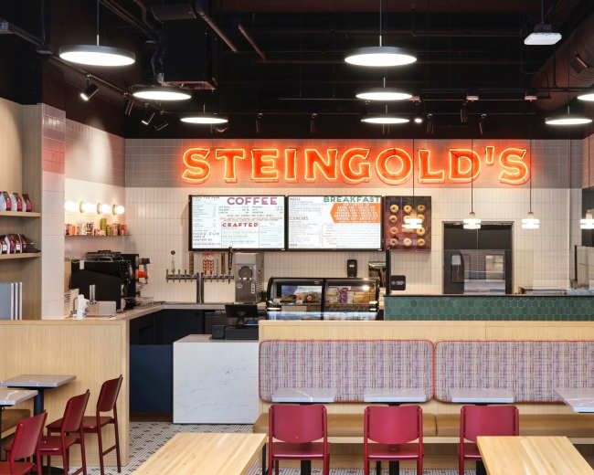 The image shows the interior of a modern café named "Steingold's," featuring a counter with a menu, seating areas with tables and red chairs, and neon signage.