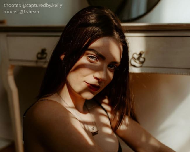 A woman sits beside a dresser, partially illuminated by sunlight filtering through window blinds, casting shadows across her face and arms.