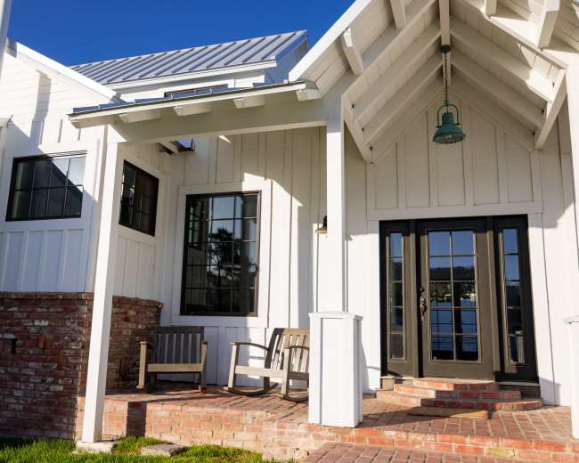 A white house with a brick porch and two wooden chairs under a triangular roof overhang.