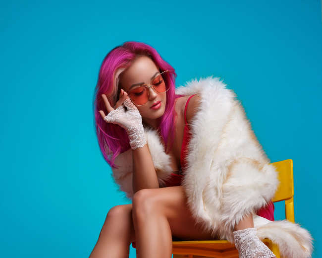 A woman with pink hair sits on a yellow chair, wearing a red outfit and high-heeled sandals, against a bright blue and pink background.