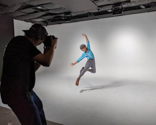 A child wearing a blue shirt and gray pants jumps in a photography studio while a photographer captures the moment.