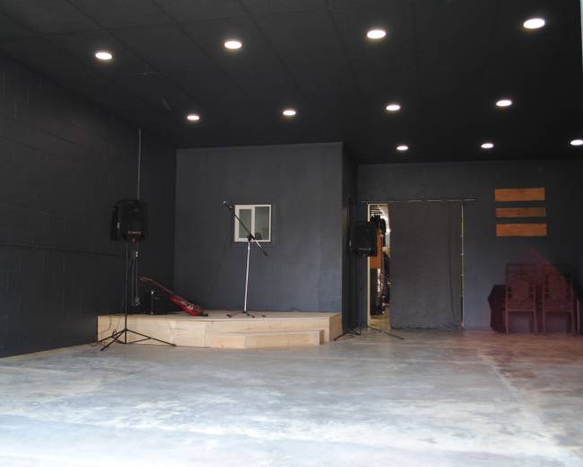 The image shows a dimly lit performance space with a raised wooden stage, a microphone stand, and speakers on either side, leading to an entrance in the background.