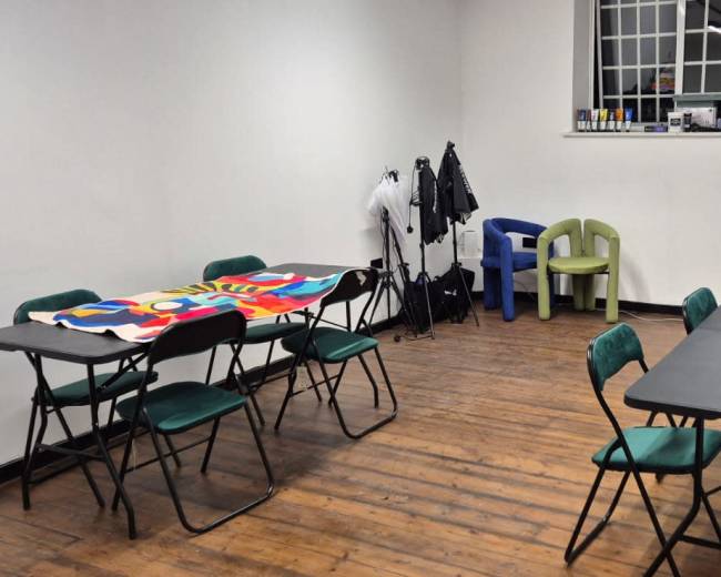 A spacious, empty room features two sets of folding tables surrounded by chairs, with a colorful cloth draped over one table and a small clothing rack against the wall.