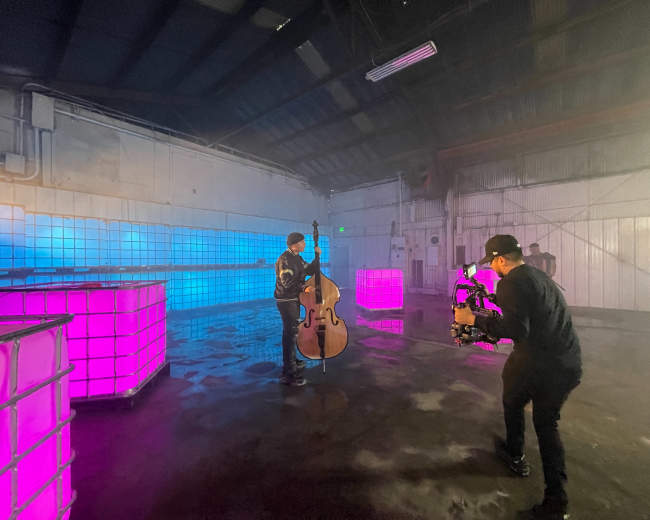 A musician plays a double bass in a foggy, industrial space with brightly lit cube structures.