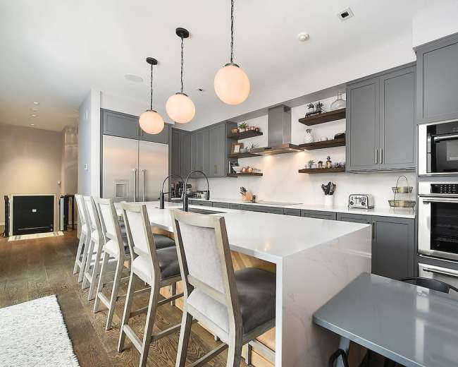 A modern kitchen features gray cabinetry, a large white island with bar seating, and pendant lights hung above.