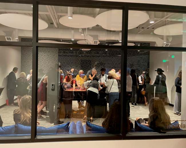 A group of people gathers around a table in a well-lit meeting room, visible through large windows.