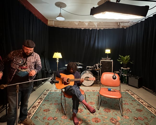 A musician sits on a chair playing an acoustic guitar in a dimly lit room with black curtains, while another person stands nearby with a microphone.