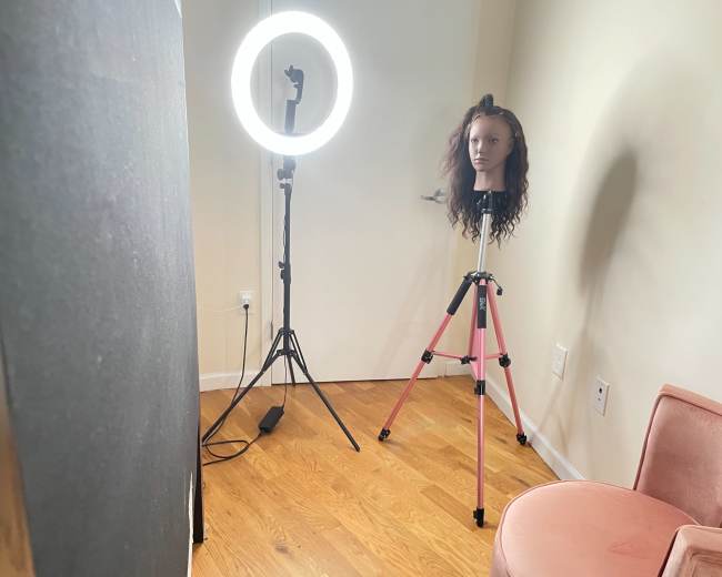 A mannequin head with curly hair sits on a pink tripod near a ring light in a room with wooden flooring.