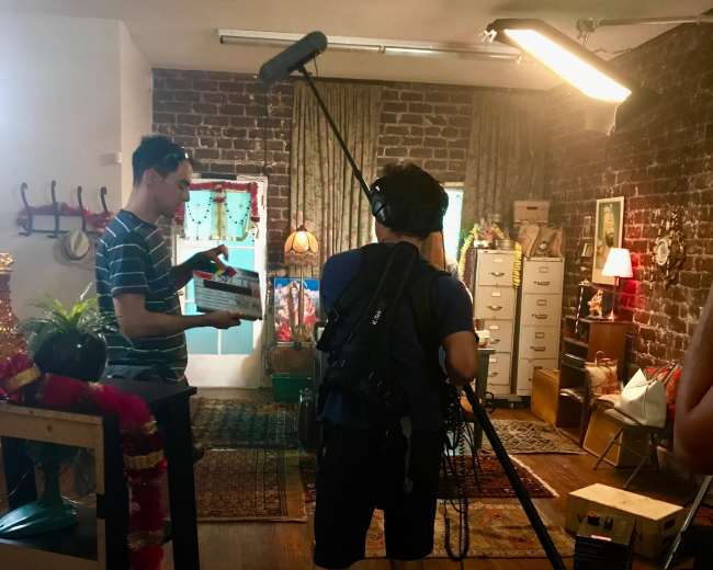 A small film set features two crew members, one holding a clipboard and the other using a boom microphone, amidst a room decorated with vintage furnishings and colorful curtains.
