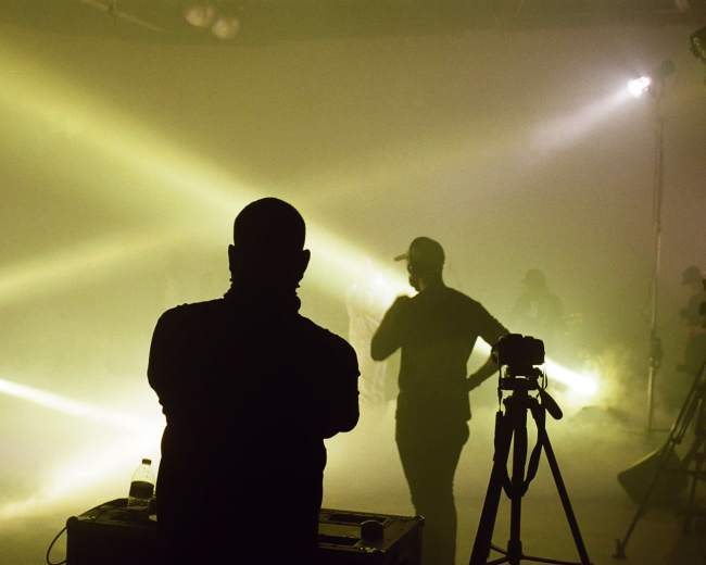 A silhouetted figure stands in front of stage lights while others perform in the background.