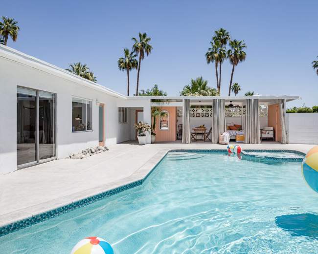 A swimming pool with colorful beach balls is surrounded by a modern house and palm trees under a clear sky.