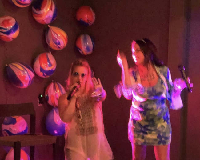 Two women are performing karaoke with microphones on a stage decorated with colorful balls and a spotlight.