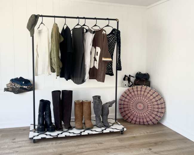 A metal clothing rack displays a variety of garments alongside several pairs of boots and a decorative round rug on a wooden floor.