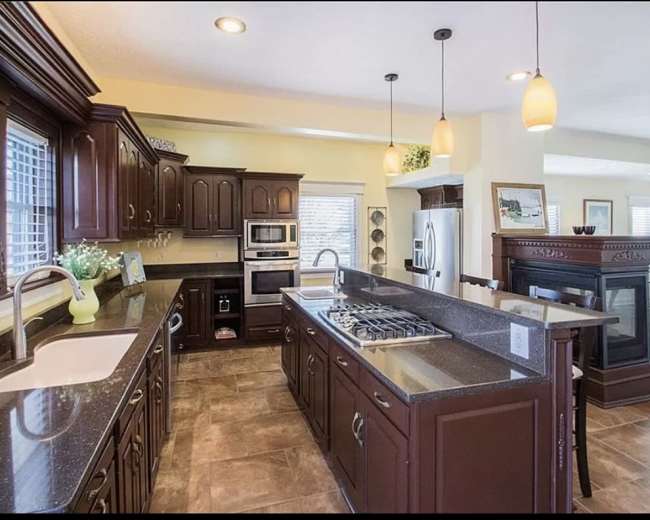 The image shows a spacious kitchen featuring dark wooden cabinets, stainless steel appliances, and a large countertop with a gas stove.