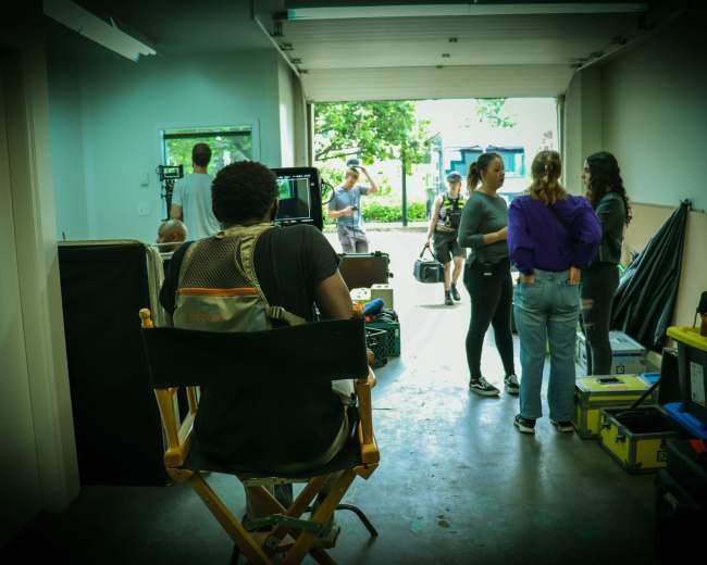 A film crew prepares for a shoot in a garage-like space, with a director seated in a chair facing the action outside.