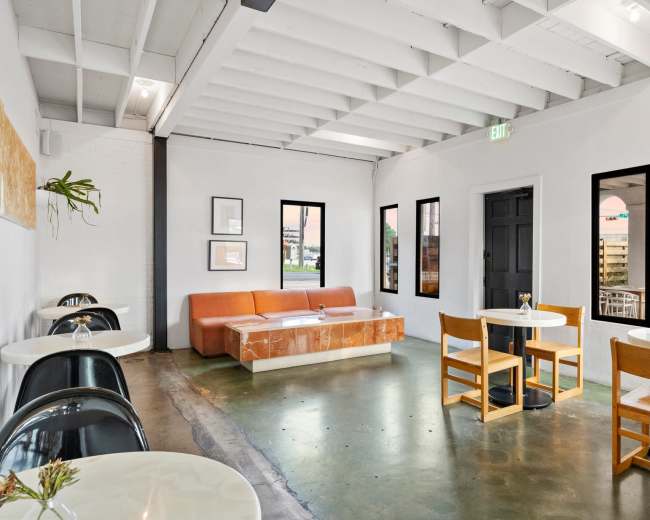 The image shows a modern cafe interior with white walls, wooden furniture, and a large orange couch.