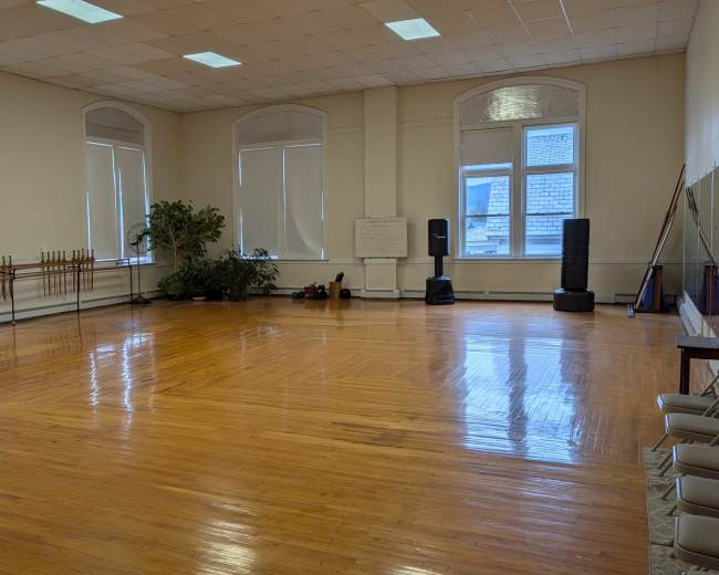 The image shows a spacious, empty dance studio with wooden flooring, large windows, and mirrors along the walls.