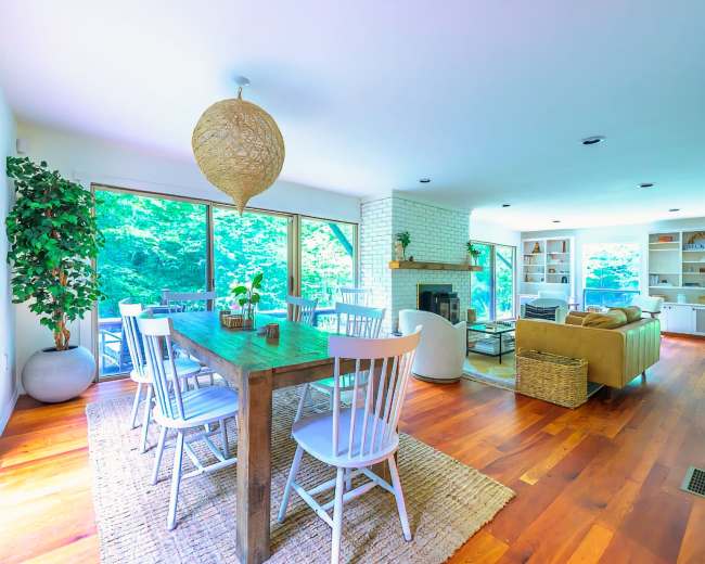A bright, open living space featuring a dining table with white chairs, large windows overlooking greenery, and a cozy seating area with a sofa and fireplace.