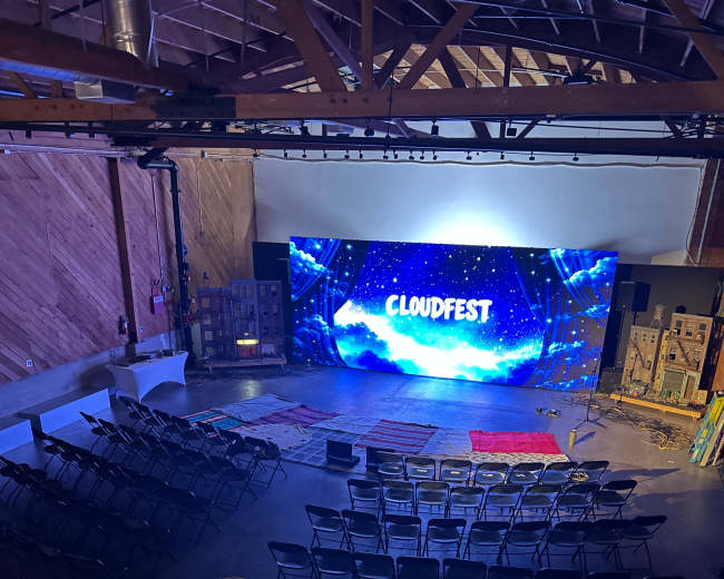 The image shows an indoor event space with rows of chairs arranged in front of a large screen that displays "CLOUDFEST."