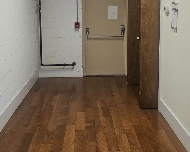 The image shows a corridor with wooden flooring leading to a closed door at the end, illuminated by fluorescent lights above.