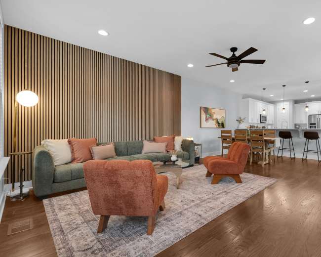 The image shows a modern living room featuring a green sofa, orange accent chairs, and a dining area with high chairs and a kitchen in the background.