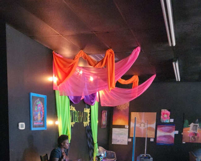 A brightly decorated indoor space features a vendor booth with colorful fabric drapes, artwork on the walls, and a person seated at a table with various crafts.