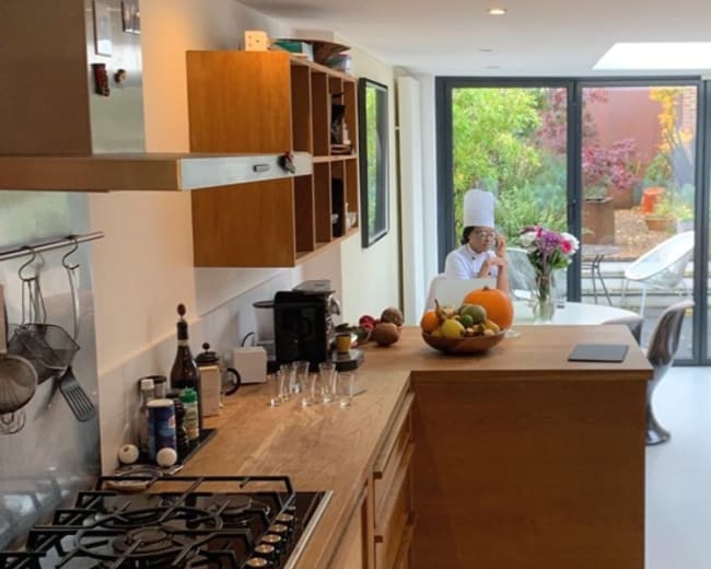 A modern kitchen opens up to a bright dining area with a view of a garden, featuring a person seated at a table surrounded by plants and natural light.