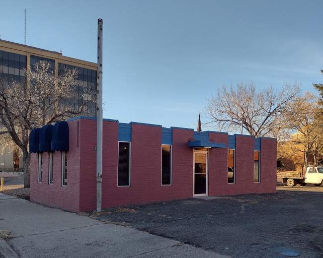 The image shows a small, pink building with blue accents and several windows, situated on a quiet street near a parking lot.