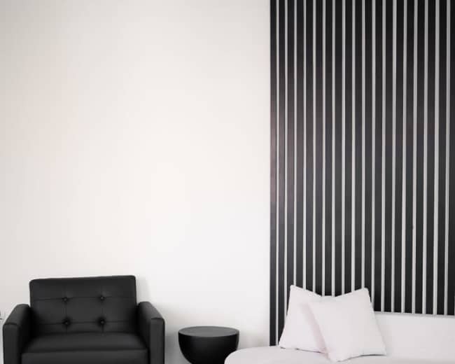 A black armchair and a white, curved sofa are positioned against a striped wall in a minimalist room with light wooden flooring.