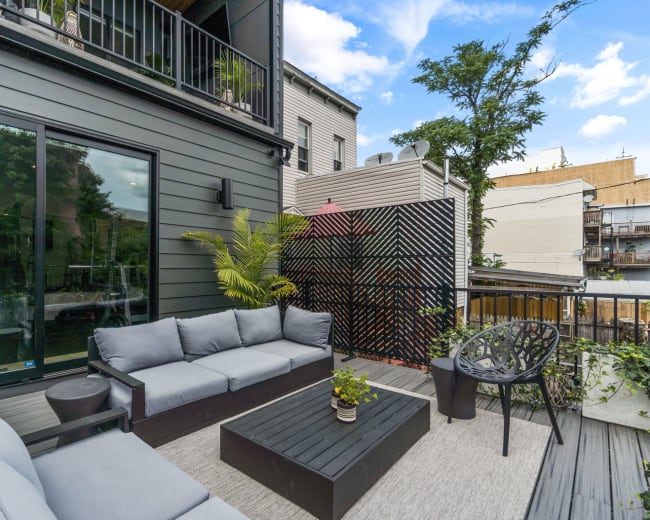 A modern outdoor patio features a gray sectional sofa, a black coffee table, and decorative greenery surrounded by buildings and blue sky.