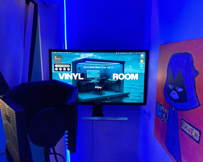 A computer monitor displays a vibrant graphic of a music studio setup labeled "VINYL ROOM," with ambient blue lighting and a colorful artwork on the wall nearby.