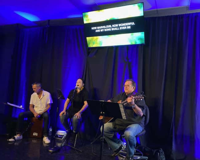 Three musicians perform on stage with a microphone and instruments, while song lyrics are displayed on a screen behind them.