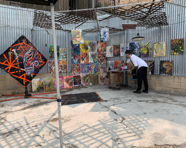 An artist is working on a table in a creative outdoor space surrounded by colorful paintings displayed on the walls.