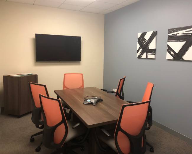 The image shows a modern conference room with a wooden table surrounded by six orange and black chairs, a wall-mounted television, and two abstract paintings.