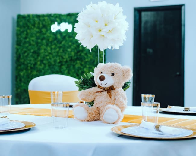 A round table is set with yellow and white decorations, featuring a teddy bear at the center and a large flower arrangement.