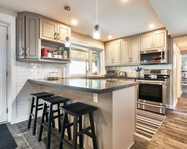 The image shows a modern kitchen featuring a central island with three black stools, stainless steel appliances, and light gray cabinets.