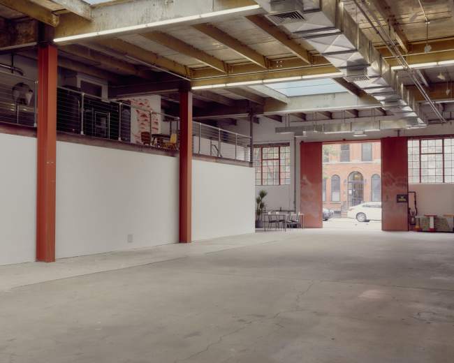 The image shows an empty industrial space with exposed beams and large windows, leading to an exterior view of a street and buildings.