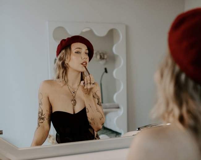 A woman with tattoos is applying lipstick while looking into a mirror, wearing a black top and a red beret.