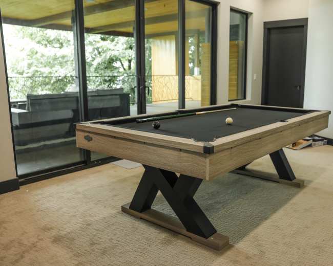 A modern pool table with a black felt surface is positioned in a room with large windows and neutral-colored walls.