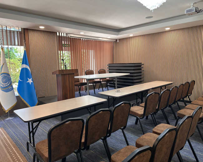 The image shows a conference room set up with several rows of chairs facing a table, flanked by two flags and a podium at the front.