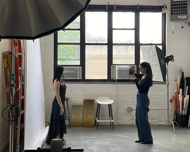 A photographer captures a model in a green dress in a well-lit studio with large windows and various backdrop materials.