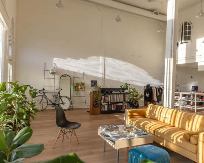 The image shows a spacious room with large windows, a yellow sofa, a coffee table filled with magazines, and various plants, along with a bicycle and shelves in the background.