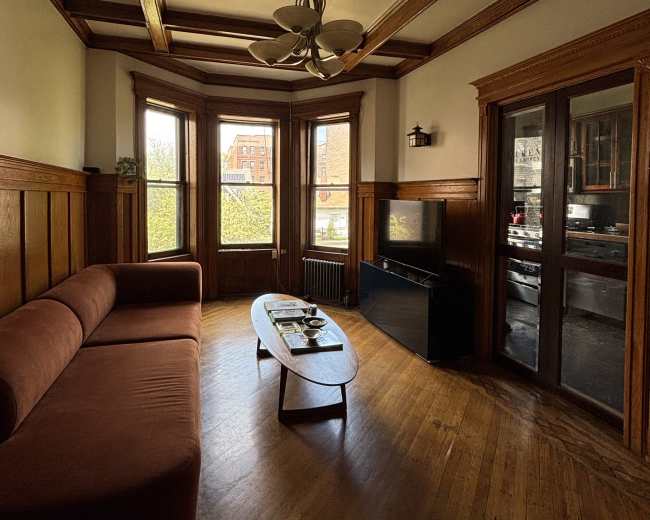 The image depicts a furnished living room with a brown sofa, a coffee table, and large windows allowing natural light to illuminate the wooden paneling and flooring.